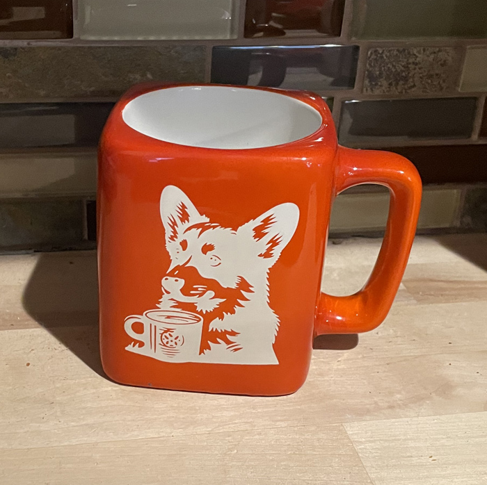 Orange ceramic coffee mug with a corgi drinking coffee engraved on the front. Corgi Mug